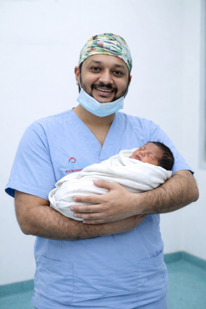 The joy of new life is beautifully captured in this moment as Dr. Jignesh Gala, dressed in scrubs and a colorful surgical cap, cradles a newborn baby wrapped in a soft white blanket. The serene hospital setting underscores the dedication of healthcare professionals who guide families through the miracle of childbirth. This image reflects not only the arrival of a new life but also the compassion and commitment of doctors who stand at the heart of neonatal care.