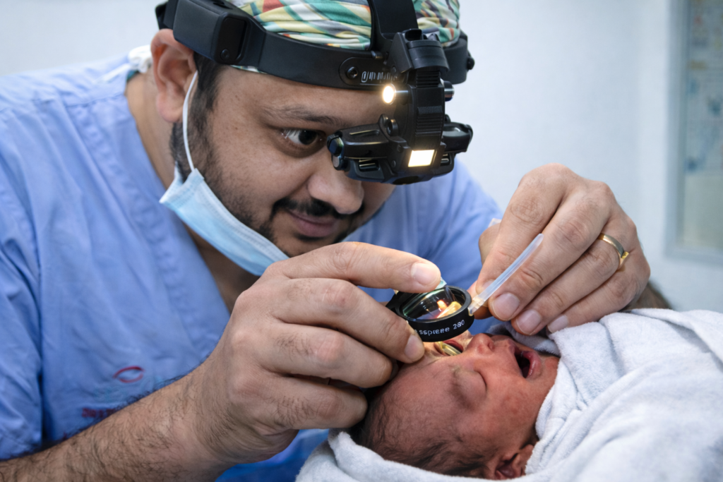 An ophthalmologist DR Jignesh Gala wearing surgical scrubs and an indirect ophthalmoscope examines a premature newborn’s eye for retinopathy of prematurity (ROP), using a handheld lens while the baby is swaddled in a white blanket in a clinical setting.