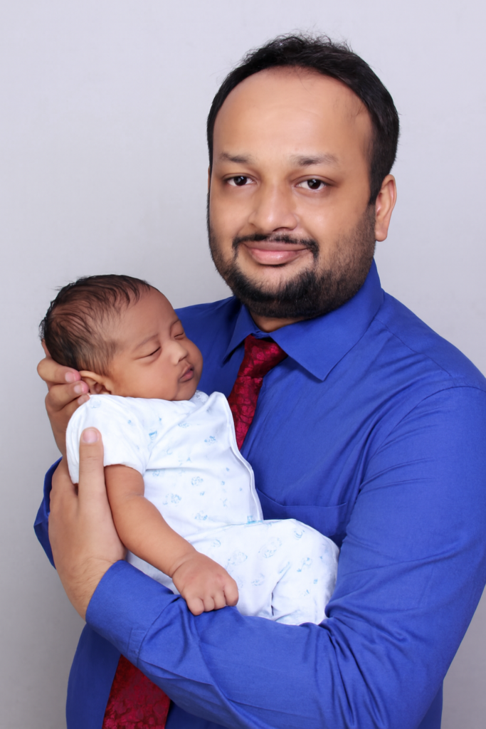 DR Jignesh Gala Holding Newborn Baby in Studio Portrait Dr Jignesh Gala in a blue shirt and red tie faces the camera while gently holding a sleeping newborn baby dressed in white, against a plain background.